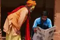 Ein Mann liest eine Zeitung, während ein Sadhu in traditioneller Kleidung ihm über die Schulter blickt. Die Szene spielt vermutlich in Nepal oder Indien und zeigt eine Begegnung zwischen traditionellem und modernem Leben.