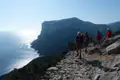 Wanderer mit Wanderstöcken auf einem steinigen Küstenpfad. Das Meer und eine steile Klippe sind im Hintergrund sichtbar.