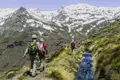 Wanderer auf einem Bergpfad mit schneebedeckten Gipfeln im Hintergrund. Ein Bach begleitet den Weg.
