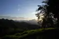 Sonnenuntergang hinter den Bergen, gesehen von einem Aussichtspunkt mit Gras und einem Baum im Vordergrund.