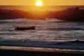 Sonnenuntergang über dem Meer mit einem teilweise zerstörten Steg im Vordergrund und Menschen, die auf einer Plattform den Sonnenuntergang beobachten.