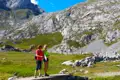 Zwei Personen wandern in einer bergigen Landschaft und stehen auf einer Brücke. Im Hintergrund sind steile Felswände und grüne Wiesen zu sehen.