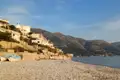 Blick auf einen Kiesstrand mit Booten, Häusern am Hang und Bergen im Hintergrund.