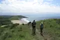 Wanderer mit Rucksäcken auf einem Küstenpfad mit Blick auf den Strand und das Meer.