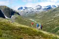 Drei Wanderer mit Rucksäcken besteigen einen grünen Berghang. Im Hintergrund erstreckt sich ein Tal mit einem Fluss und schneebedeckten Bergen.