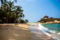 Palmengesäumter Sandstrand mit einer Hütte auf einer Anhöhe und türkisblauem Meer.