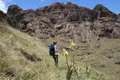 Wanderin mit Rucksack und Wanderstöcken pausiert auf einem Bergpfad neben Wildblumen und betrachtet die felsige Landschaft.