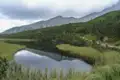 Zwei Wanderer auf einem Bergpfad neben einem See, umgeben von bewaldeten Hängen und Bergen.