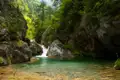 Ein kleiner Wasserfall ergießt sich in einen klaren, türkisfarbenen Pool inmitten einer bewaldeten Schlucht.