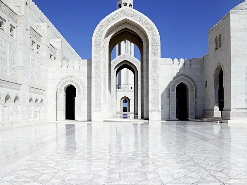 Blick auf einen der Innenhöfe der Sultan Qaboos-Moschee in Maskat mi tweiß- glänzendem Marmorboden. Durch einen spitzen Torbogen erblickt man einen Teil des Minaretts.