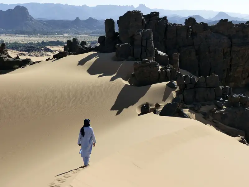 Person durchquert eine Sanddüne in einer bergigen Wüstenlandschaft.