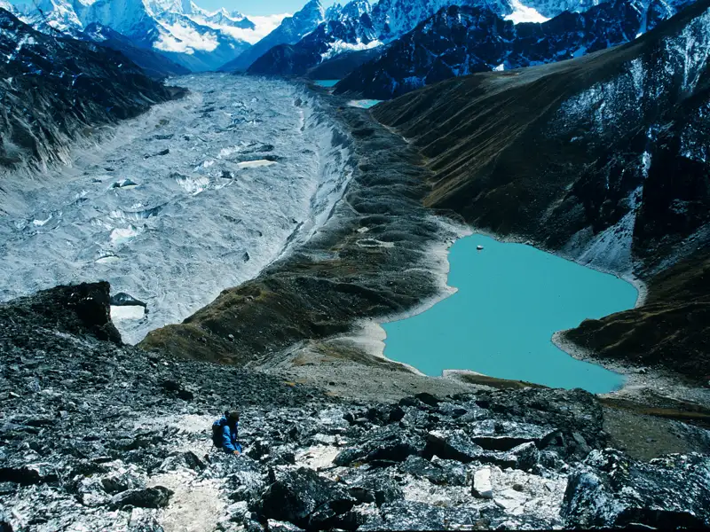 Gletscherlandschaft im Himalaya mit einem türkisfarbenen See. Ein Wanderer befindet sich im Vordergrund und erkundet die Umgebung.