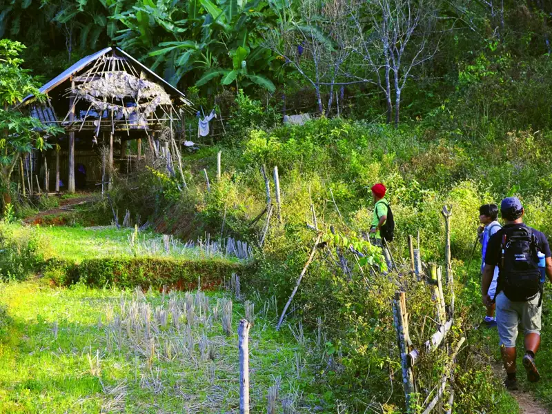 Drei Wanderer auf einem Pfad, der an einer Hütte und Feldern in ländlicher Umgebung vorbeiführt.