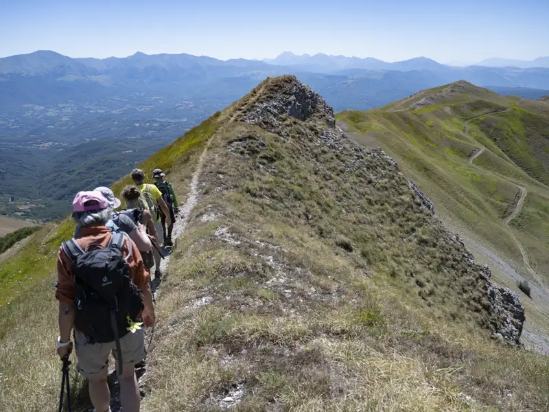 Wanderer mit Rucksäcken und Wanderstöcken auf einem schmalen, grasbewachsenen Berggrat. Im Hintergrund sind weitere Berge und Täler zu sehen.
