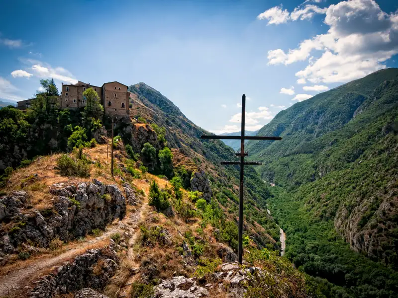 Panoramablick auf ein Bergdorf und ein grünes Tal mit einem Kreuz im Vordergrund.