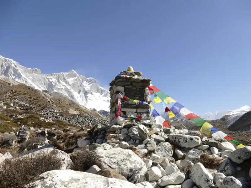 Ein Steinmal mit bunten Gebetsfahnen markiert einen Punkt im Himalaya, mit den schneebedeckten Gipfeln im Hintergrund.