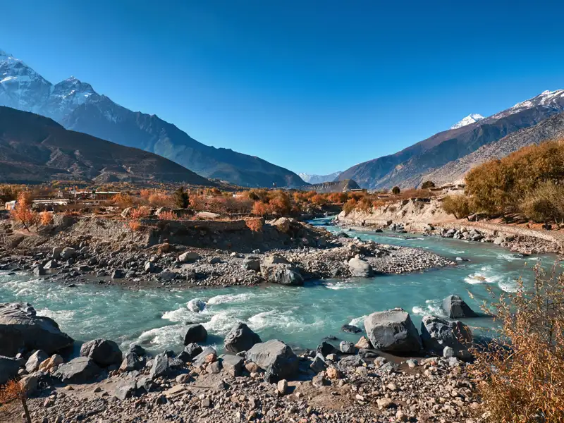 Panoramablick auf einen Fluss, der durch ein Gebirgstal fließt. Die Landschaft zeigt herbstliche Bäume und schneebedeckte Gipfel.