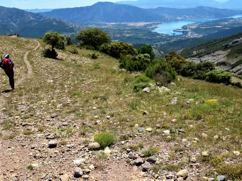 Wanderer auf einem steinigen Bergpfad mit Blick auf einen See und Berglandschaft im Hintergrund.