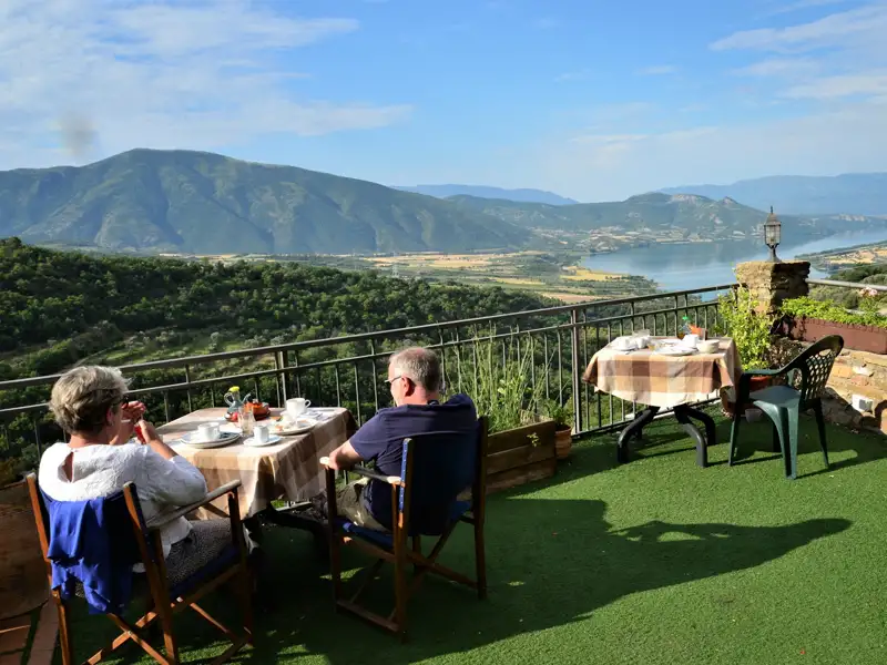 Zwei Personen frühstücken auf einer Terrasse mit Blick auf einen See und Berge im Hintergrund.