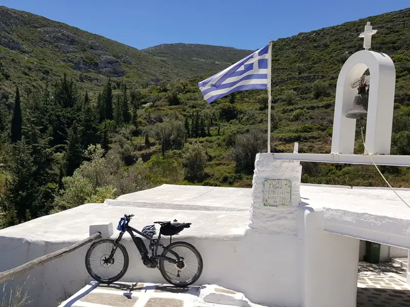 E-Bike vor einer weißen Kapelle in den griechischen Bergen. Eine griechische Flagge ist zu sehen.