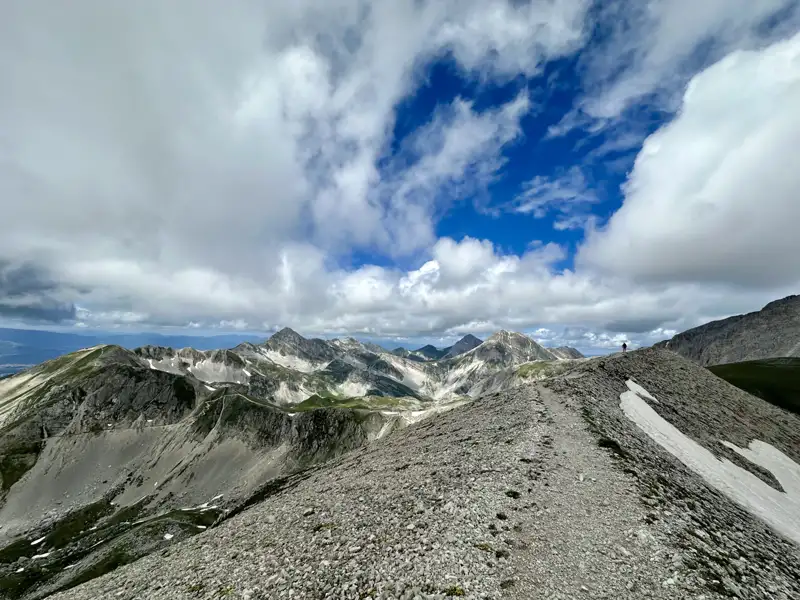 Panoramablick von einem Berggipfel auf eine Bergkette mit einem felsigen Pfad und Schneefeldern.