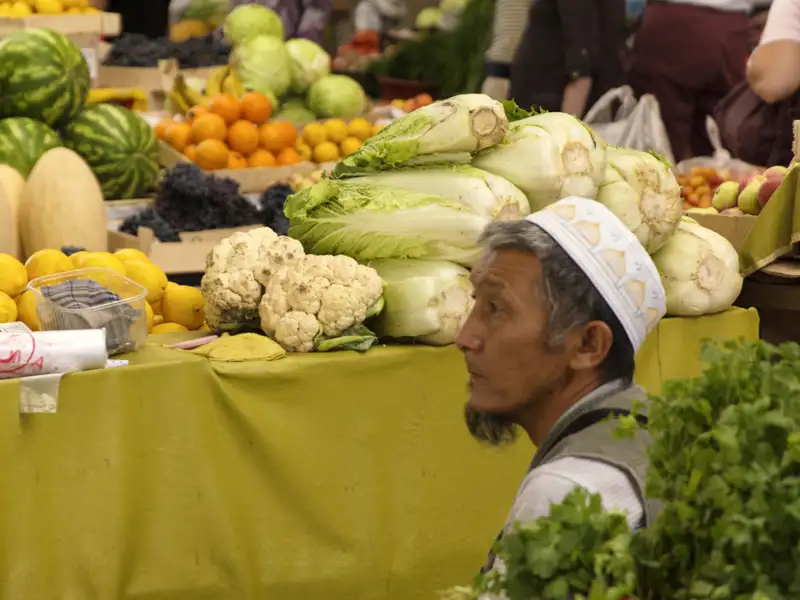 Mann an einem Marktstand mit Chinakohl, Blumenkohl, Zitronen, Melonen und weiterem Obst und Gemüse.