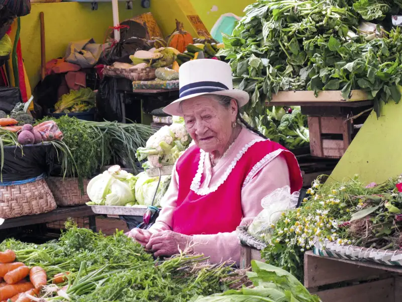 Marktfrau mit Hut verkauft frisches Gemüse und Kräuter.
