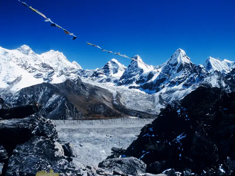 Gebetsfahnen wehen im Wind vor der Kulisse schneebedeckter Himalaya-Gipfel und eines Gletschers.