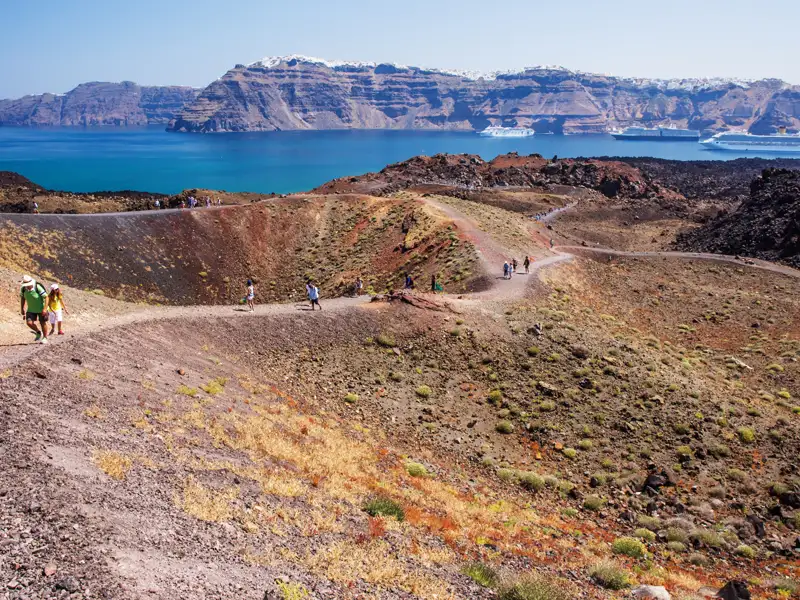 Wanderer auf einem vulkanischen Pfad mit Blick auf die Caldera und Kreuzfahrtschiffe im Hintergrund.