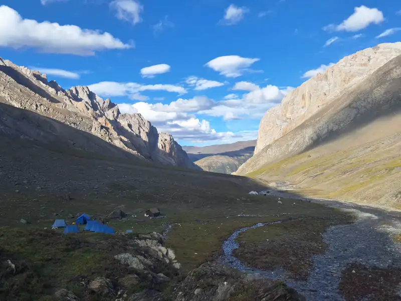 Basislager in einem Gebirgstal, umgeben von hohen Bergen.  Blaue Zelte und ein kleiner Fluss sind sichtbar.