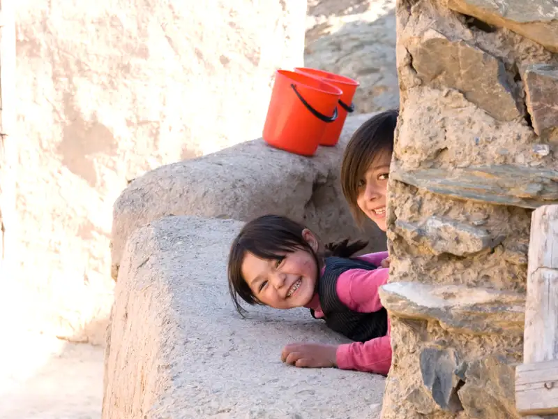 Zwei Kinder, die sich hinter einer Mauer verstecken, lächeln in die Kamera.  Zwei rote Eimer stehen auf der Mauer.