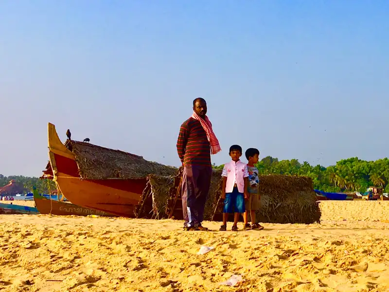 Ein Mann und zwei Kinder stehen am Strand neben einem traditionellen Fischerboot, das möglicherweise für Fischfang oder Bootsfahrten genutzt wird.