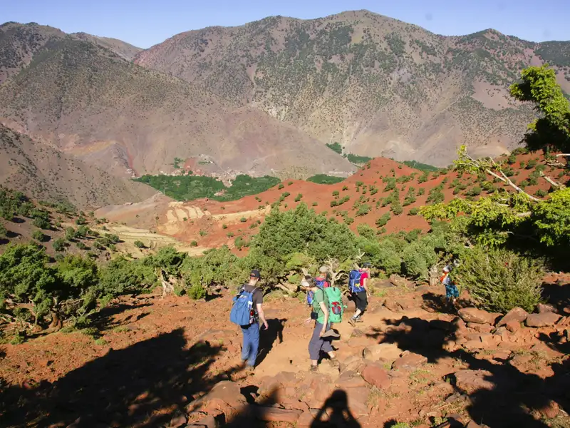 Wandergruppe auf einem Bergpfad im Atlasgebirge. Roter Boden und grüne Vegetation prägen die Landschaft.
