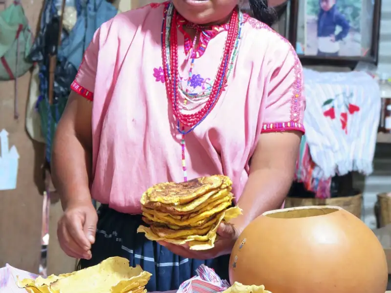 Frau in traditioneller Kleidung präsentiert einen Stapel frisch zubereiteter Tortillas.