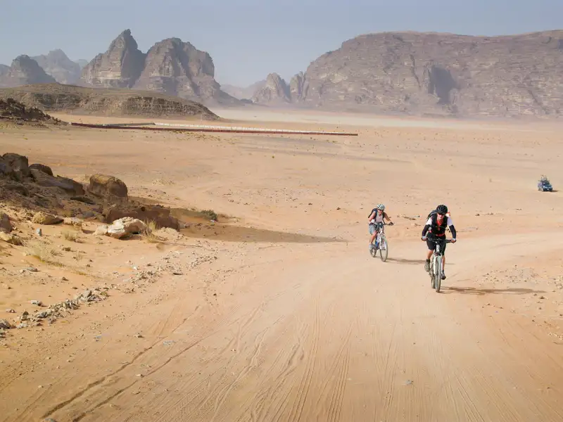 Zwei Mountainbiker fahren auf einem sandigen Weg in der Wüste. Im Hintergrund sind Felsformationen zu sehen.