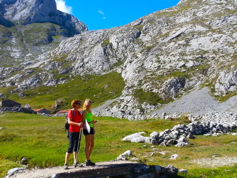 Zwei Personen wandern in einer bergigen Landschaft und stehen auf einer Brücke. Im Hintergrund sind steile Felswände und grüne Wiesen zu sehen.