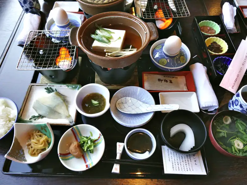 Traditionelles japanisches Frühstück mit verschiedenen Speisen wie Fisch, Tofu, Suppe, Reis und Beilagen.